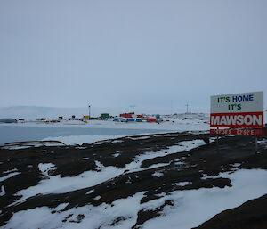 Mawson station with fresh snowfall all around and the station sign reading “It’s home, it’s Mawson” in the foreground