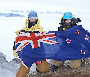 Two expeditioners hold up a New Zealand flag while crouching on the top of Mt Henderson