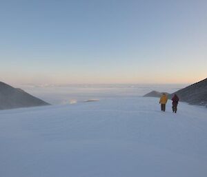 Two expeditioners walk in a snow drift, elevated and surrounded by landscape consisting of rocks and more snow