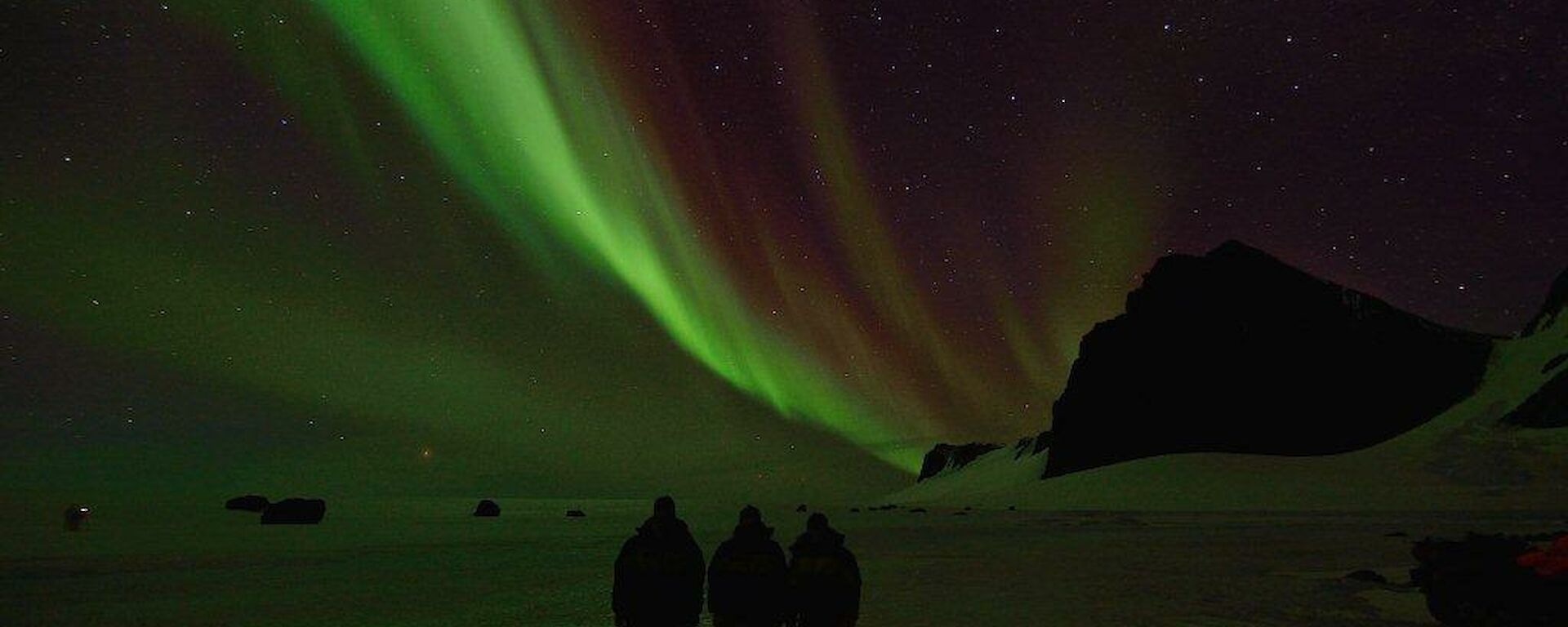 Three expeditioners standing on the ice with an Aurora over the silhouette of the North Masson range