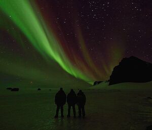 Three expeditioners standing on the ice with an Aurora over the silhouette of the North Masson range