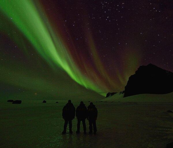 Three expeditioners standing on the ice with an Aurora over the silhouette of the North Masson range