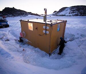 An expeditioner shovels snow away from the side of Colbeck Hut