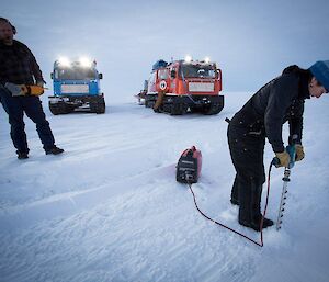 An expeditioner drills the sea ice while another expeditioner stands by holding a rescue throw bag
