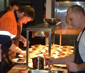 The chef and one expeditioner preparing the main course of eye fillet to be served up in the kitchen, with the plates under heating lamps