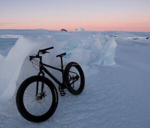 A bicycle lays on its side against a small snow drift, with a beautiful sunset sky in the background