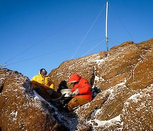 Two expeditioners perched between rocks and out of the wind whilst installing the channel radio repeater on top of Mt Henderson