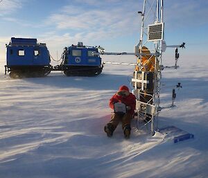 An expeditioner sitting next to the AWS begins the data download — with the blue Hägglunds in the background and with blowing snow and 30 knots of wind