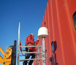 Andy standing in a cage and being lifted by a JCB so as to inspect the BGAN antenna mount