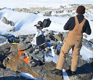 A man standing in overalls surrounded by rocks and snow, at center of image is technical equipment — a remote penguin camera