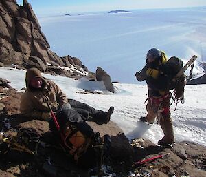 One expeditioner lazing in the sun with views out and across the Antarctic plateau behind him as another expeditioner laden with climbing gear goes through some last minute preparations before seeting off on the climb