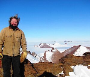 An expeditioner pauses to pose for a photograph as he stands in a sheltered saddle below the summit of Mt Hordern. Mt Dunlop can be seen in the background along with a number of unamed peaks — all with a dusting of snow them. The vast Antarctica Plateau stretches out and beyond the peaks.
