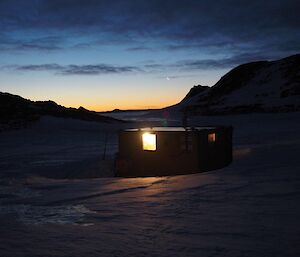 The hut pictured at dusk with the light from inside emanating out through a ting window