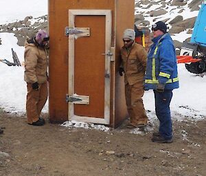 Three expeditioners stands around the hut’s toilet waiting for instructions to position it at the new hut site