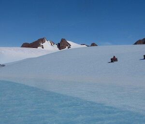 Curly and Chris riding their Quad bikes down a snow covered slope toward a frozen ice stream with the mountains in the background