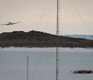 The Basler plane is seen to approach the Mawson Ski Landing Area from beyond Bechervaise Island. The ground crew consisting of the Mawson Emergency Response Team and Refueling Team along with the two passengers bound for Davis can be seen gathered on the sea ice in the foreground