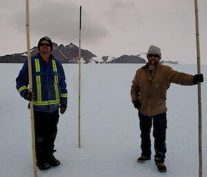 Chris and Aidan standing with three bamboo canes which mark a track junction. You can see Mt Henderson and Henderson Hut in the background