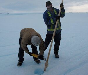 An expeditioner leans down on the ice to saw the end off a tall cane, while another expeditioner holds it upright