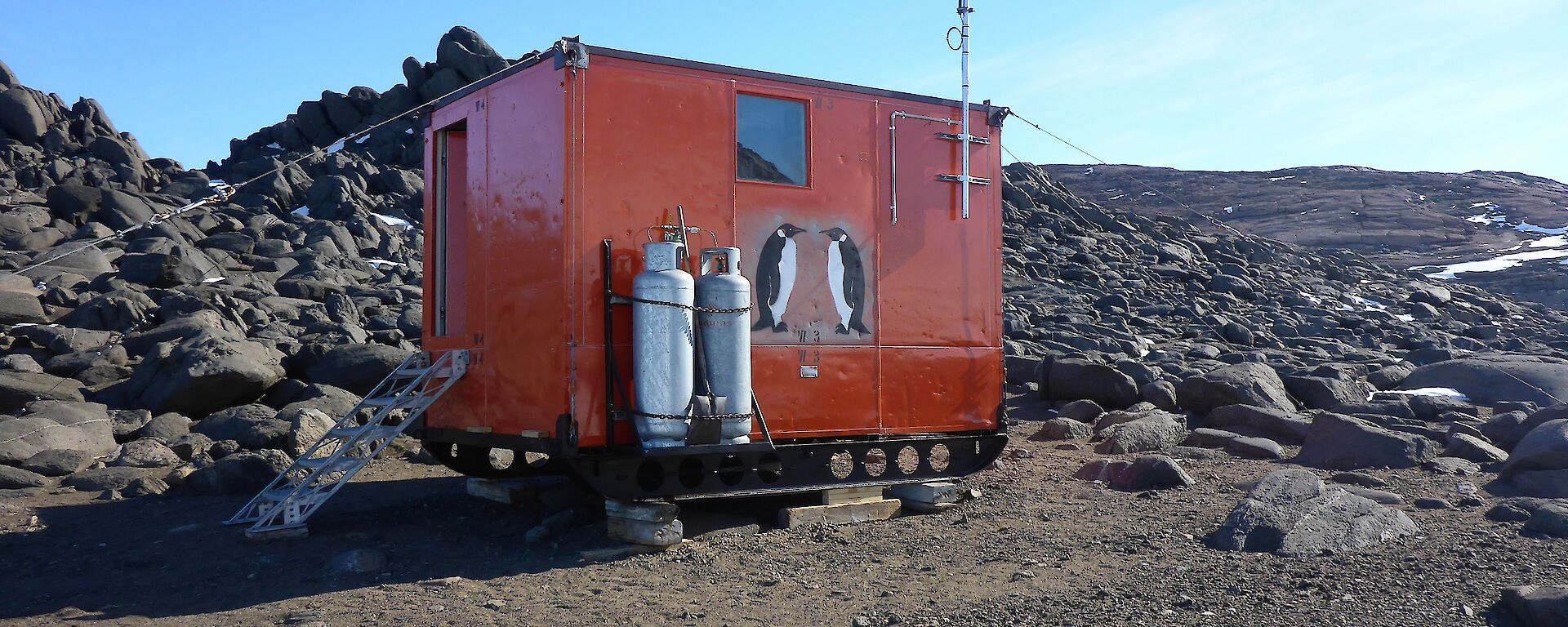 Colbeck hut sits amongst rocks. It is a brightly coloured half-sized shipping container