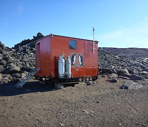 Colbeck hut sits amongst rocks. It is a brightly coloured half-sized shipping container