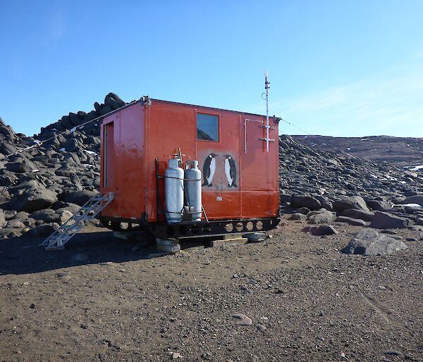 Colbeck hut sits amongst rocks. It is a brightly coloured half-sized shipping container