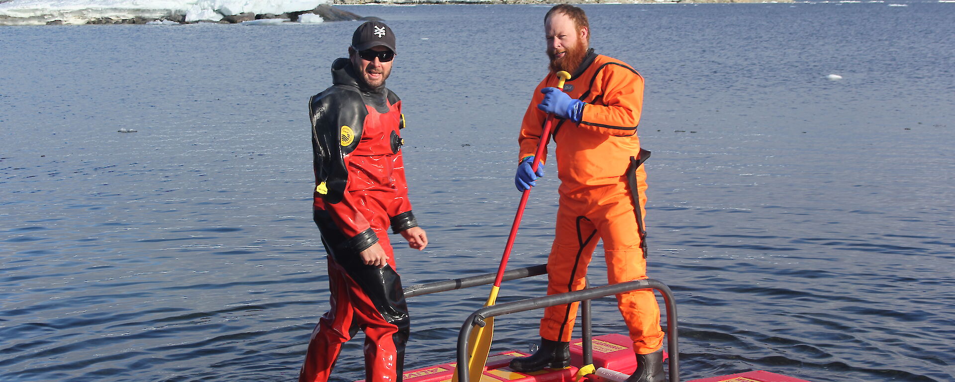 Two expeditioners wearing drysuits standing on a raft on the water