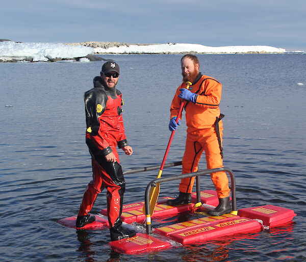 Two expeditioners wearing drysuits standing on a raft on the water
