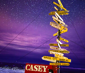 The Casey station sign, with many wooden arrows pointing to global locations, with brilliant aurora and stars lighting up the sky