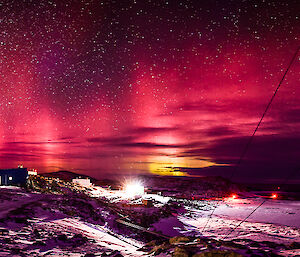 A panorama of Casey station at night with building lights on, aurora australis and stars lighting up the night sky, and the station sign in the foreground