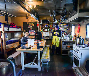 Expeditioners relax inside a small, self-contained hut with beds on left, picnic table in middle and kitchen on right