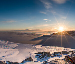 An expeditioner dressed in winter gear sits in the snow in front of a spectacular sunset