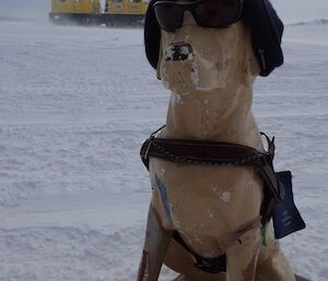 In the foreground, a plastic guide dog wearing a beanie sits on the snow covered ground, a large Hägglunds oversnow vehicle waits in the background