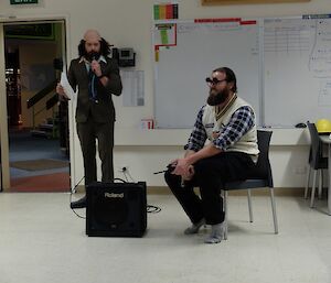 Two male expeditioners hosting quiz night, one with a faux bald patch wig and the other with beard and cricket jumper