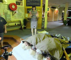The female station doctor is directing a video production where bearded men lie on their backs under a sheet to give the impression their beards have eyes.