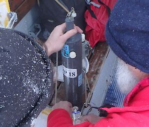 Two expeditioners hold a slim metal cylinder filled with a scientific water sample.