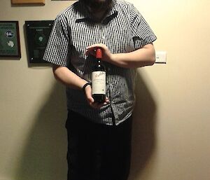 A young male expeditioner holds up a bottle of wine and smiles at the camera.