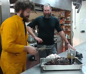 A bearded expeditioner in bright jumpsuit tends to meatloaf while another bearded male expeditioner looks on with derision.