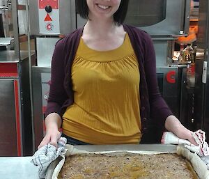 A female expeditioner with bob haircut presents a tin of cooked meatloaf and smiles.