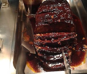 A juicy, sliced meatloaf is presented in a bain marie with knife and jug of gravy beside it. The name Hilda is written in white board marker underneath to signify who cooked it.