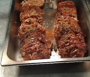 A meatloaf log has been cut up into circular patties, and sits inside a bain marie accompanied by a metal jug of gravy. The name Agnes is written on the stainless steel bench in front of the bain marie dish.