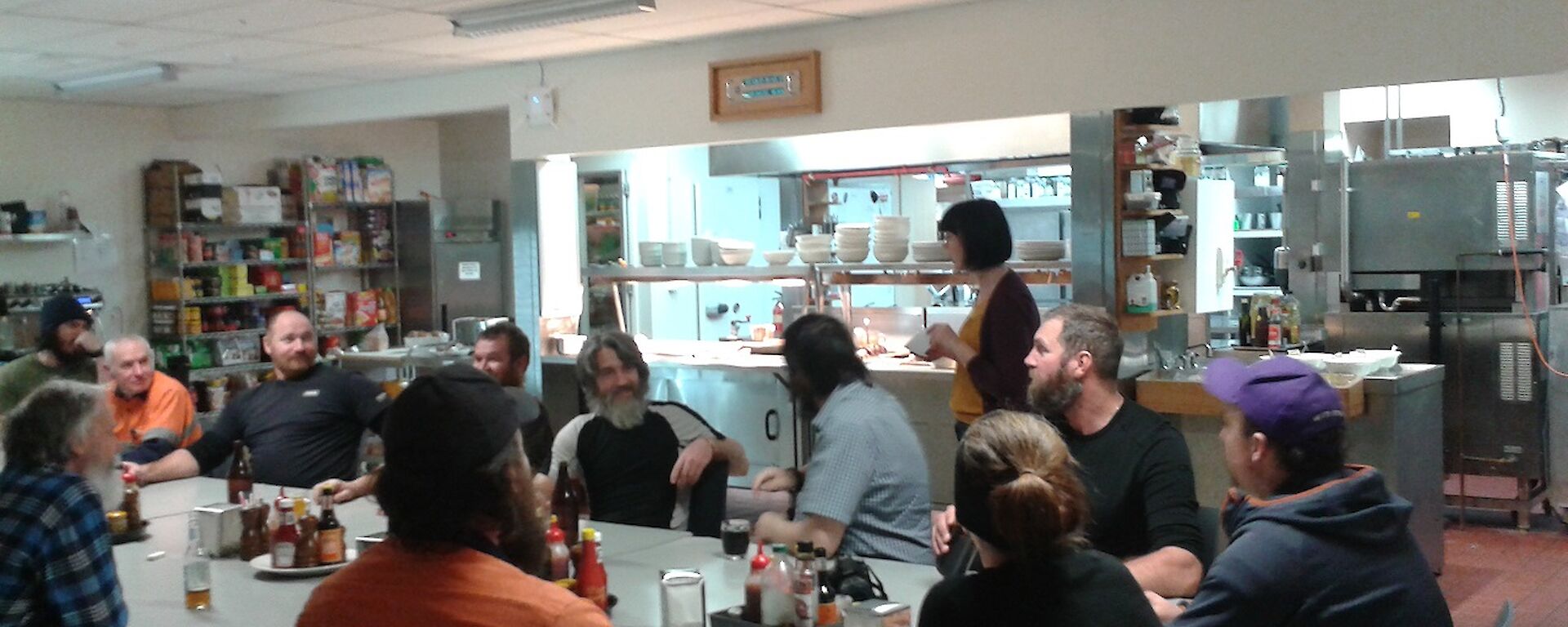 A large rectangular table is crowded with expeditioners. It sits in front of the bain marie serving area outside a kitchen. The crowd is waiting to taste several meatloaves.