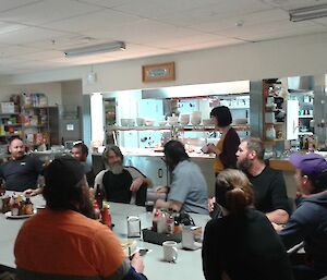 A large rectangular table is crowded with expeditioners. It sits in front of the bain marie serving area outside a kitchen. The crowd is waiting to taste several meatloaves.