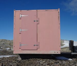 The end of a square metal hut with door and large hinges sitting amongst rocks and snow