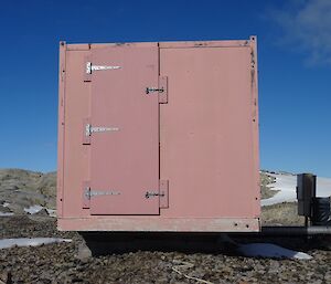 The end of a square metal hut with door and large hinges sitting amongst rocks and snow