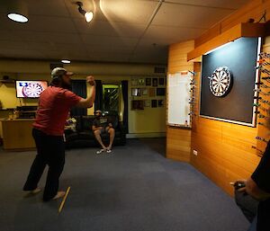 A bearded man poses to throw a dart at a dart board.