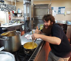 Bearded expeditioner stirs a pot with curry prawns while giving the thumbs up and looking excited.