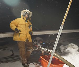 Midwinter refuelling — expeditioner leans against a pipe in full gear facing camera as snow falls around him