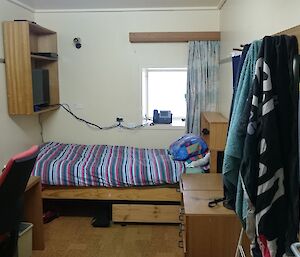 Expeditioner’s small bedroom with desk on right, bed and window against wall and clothing pegs on right.