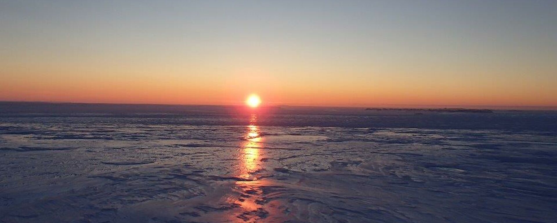 The sun sets over an icy plateau with light creating a triangular column reflecting in the slick surface