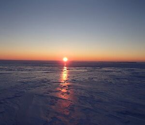 The sun sets over an icy plateau with light creating a triangular column reflecting in the slick surface